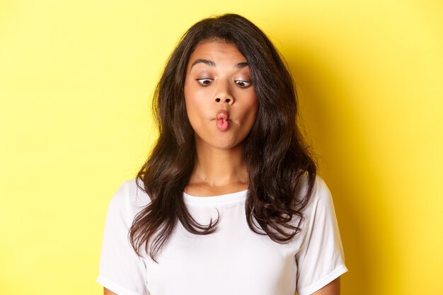 Close-up of funny african-american woman, making faces, squinting and pouting, fooling around over yellow background.