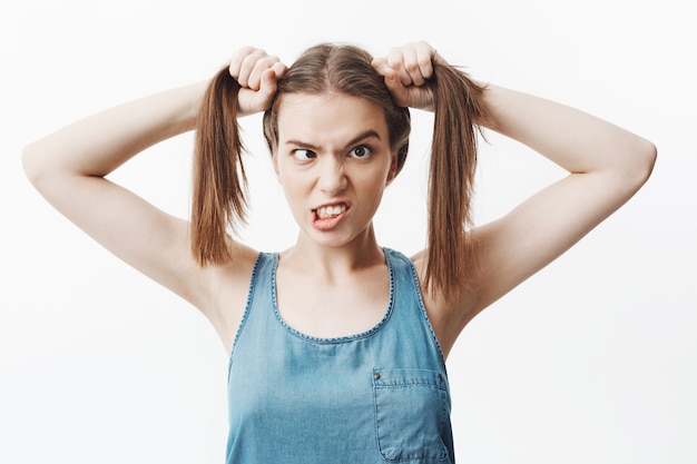 Free photo close up of funny beautiful european female student with brown hair in blue jeans shirt holding hair with both hands, mowing eyes, showing teeth,  with awkward expression, having fun.