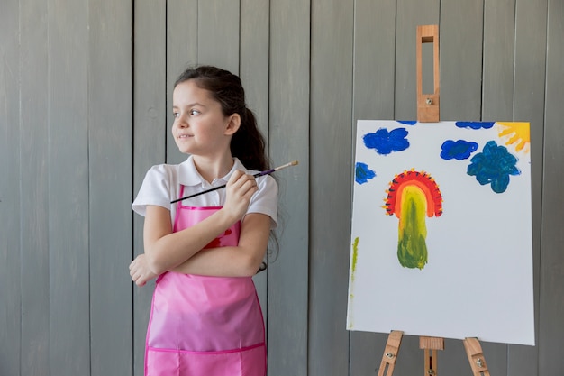 Free photo close-up of a girl holding paint brush in hand standing near the easel looking away