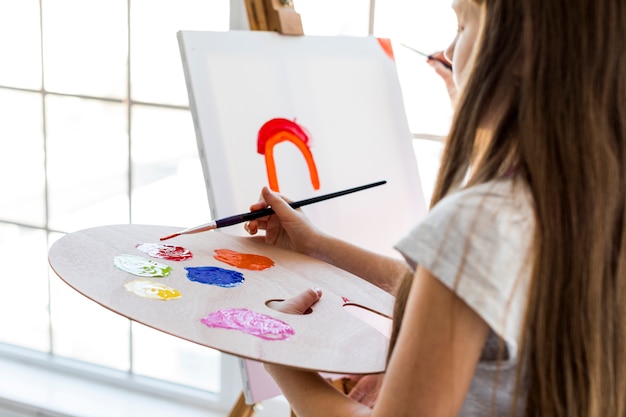 Free Photo close-up of a girl standing in front of canvas mixing the red paint with paintbrush