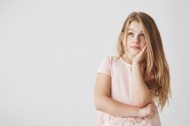 Close up of good-looking girl with blonde hair and blue eyes holding hand on cheek, looking aside with dreamy expression, thinking about holidays.