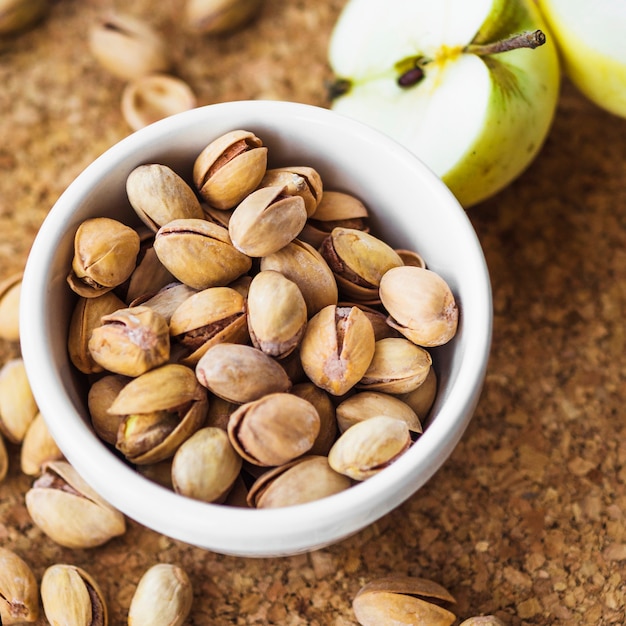 Close-up of halved apple with bowl of pistachio on cork board