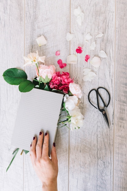 Close-up of hand holding book with hydrangea and roses flowers and scissor on wooden plank