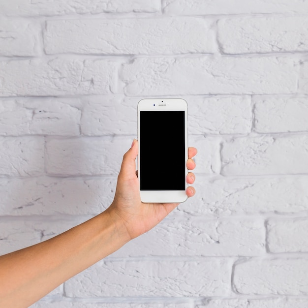 Close-up of hand holding mobile phone with blank screen in front of white wall