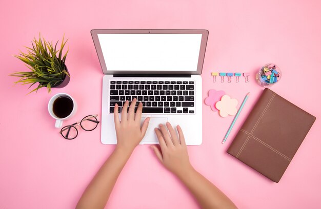 Close-up of hand typing on laptop with stationeries and coffee cup on pink backdrop