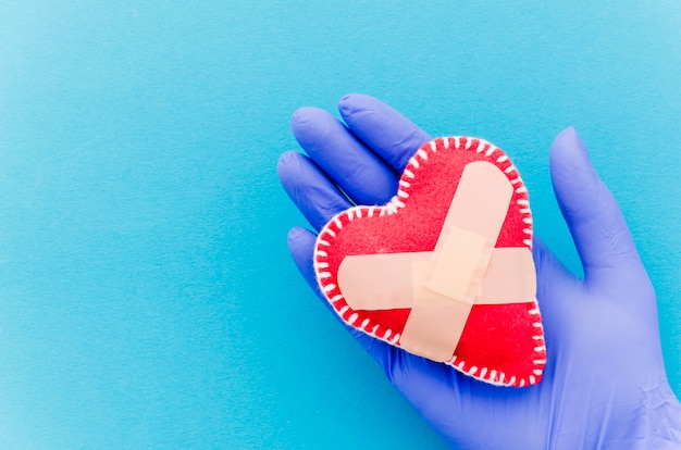 Close-up of hand wearing surgical gloves holding heart shaped stitched textile heart with crossed bandages on blue backdrop