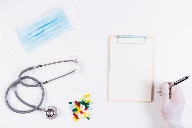 Close-up of hand writing on blank clipboard with medical equipments over white background