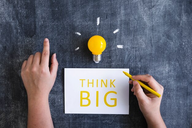 Close-up of hand writing text think big on white paper with yellow light bulb on chalkboard