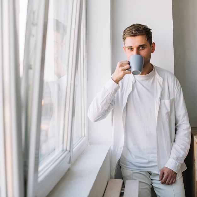 Close-up of a handsome guy drinking beverage standing near window