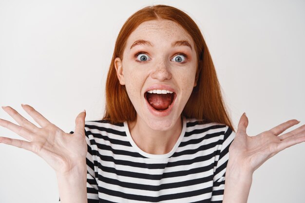 Close-up of happy redhead woman shouting from joy and happiness, looking surprised, standing over white wall