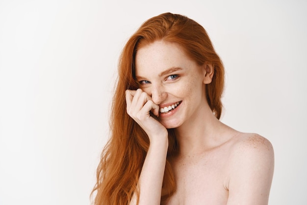 Free photo close-up of happy redhead woman with pale perfect skin, laughing and showing white teeth, standing naked on studio wall