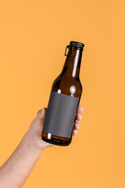 Close-up of human hand holding brown beer bottle against yellow wall backdrop