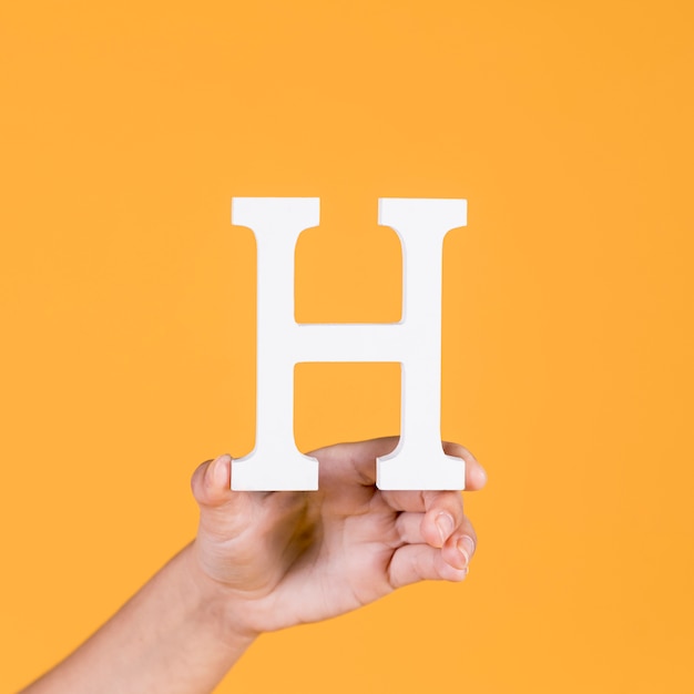 Close-up of a human hand holding up the uppercase capital letter h over yellow background