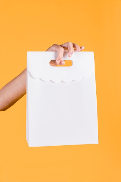 Free Photo close-up of human hand holding white paper bag on yellow wall backdrop