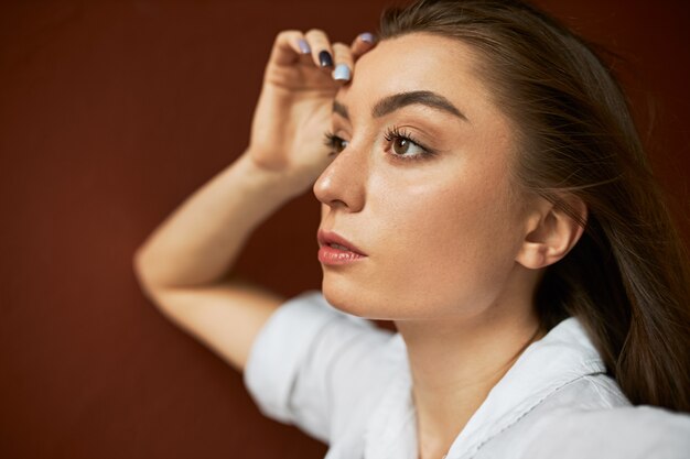 Close up image of attractive thoughtful young Caucasian woman with clean skin and loose brown hair touching face, having pensive facial expression, looking ahead of her, recollecting something