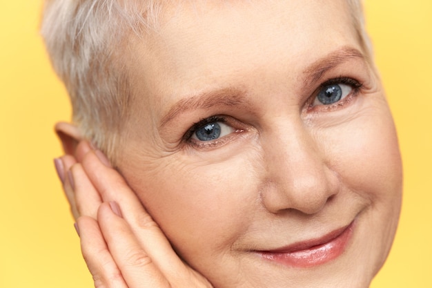 Free photo close up image of beautiful middle aged woman with blue short blonde hair and wrinkles around her blue eyes smiling, placing hands under cheek.