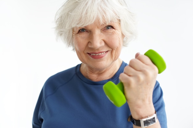 Free Photo close up image of energetic sporty mature woman with gray hair and wrinkles exercising indoors, doing bicep curls, holding green dumbbell and smiling happily. sports, age and fitness