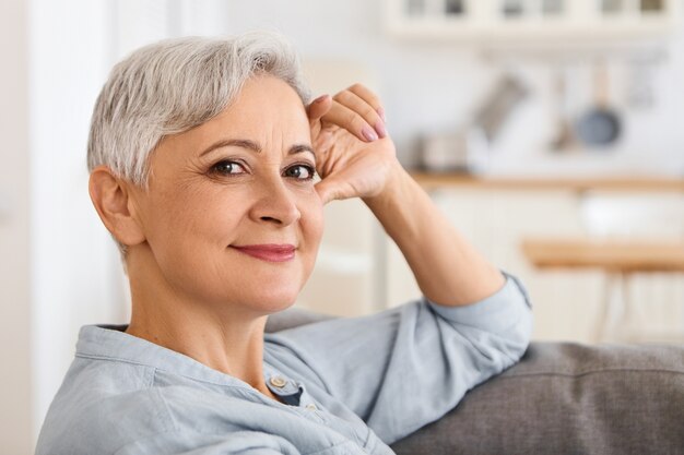 Close up image of fashionable elegant elderly woman with stylish haircut and make up relaxing indoors, sitting comfortably on couch with hand on her face, looking  with carefree smile