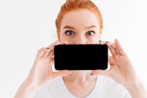 Free Photo close up image of ginger woman showing blank smartphone screen and covering her mouth