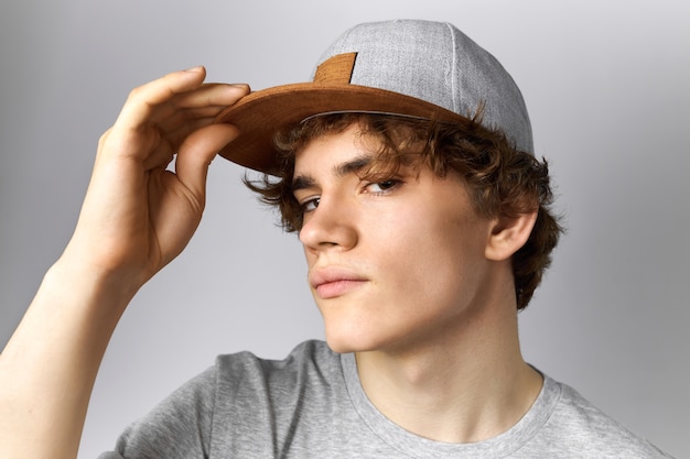 Free Photo close up image of handsome young european guy with curly hair and smooth face having fun wearing trendy snapback holding hand on peak and staring at camera with confident look. style and clothing