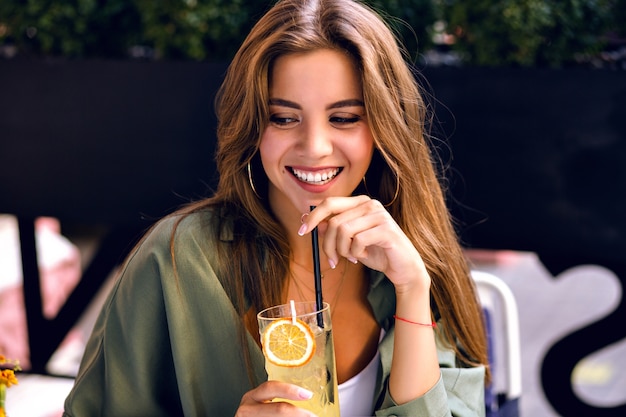 Free photo close up image of pretty brunette woman drinking tasty lemonade and looking on camera, enjoying time at city terrace, warm summertime, natural beauty.