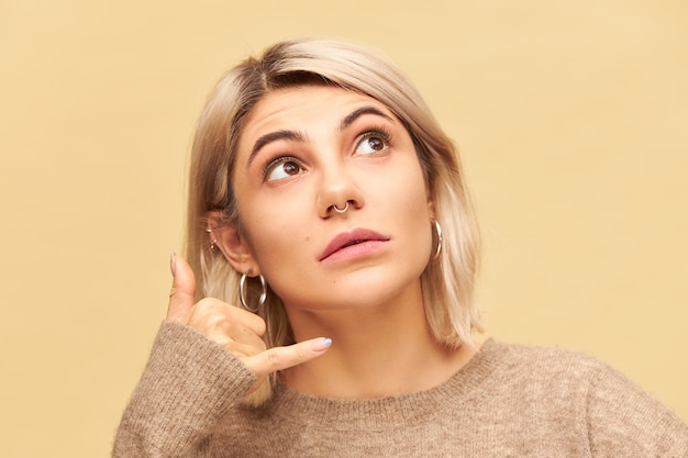 Close up image of pretty female with blonde bob hairstyle and nose ring looking up, holding hand at her ear with thumb and little finger spread widely, making gesture Call me. Body language