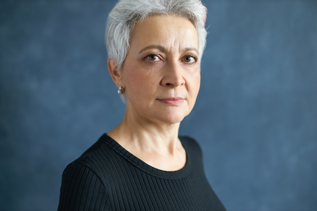 Close up image of serious attractive mature female with stylish short haircut looking at camera, frowning eyebrows posing against blank studio wall background with copy space