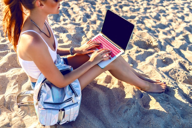 Free photo close up image of young woman sitting on the beach and working on her laptop, backpack, freelance style. work on vacation.