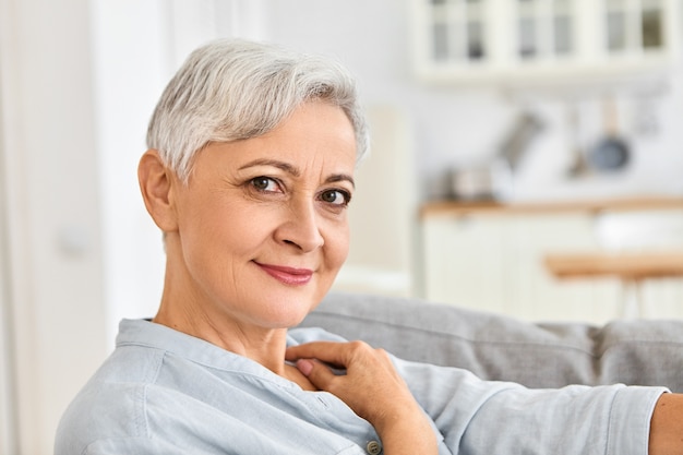 Close up indoor picture of elegant elderly woman pensioner relaxing at home sitting comfortably on couch after shower, wearing cozy cotton bathrobe with cute charming smile