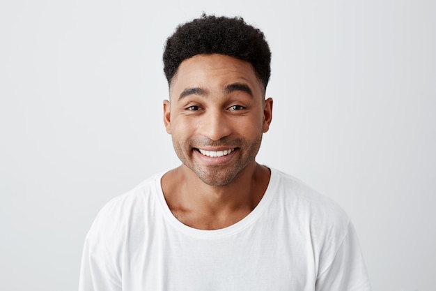 Close up isolated portrait of cheerful happy young man with afro hairstyle in casual white t-shirt smiling brightly, looking in camera with excited and joyful expression.