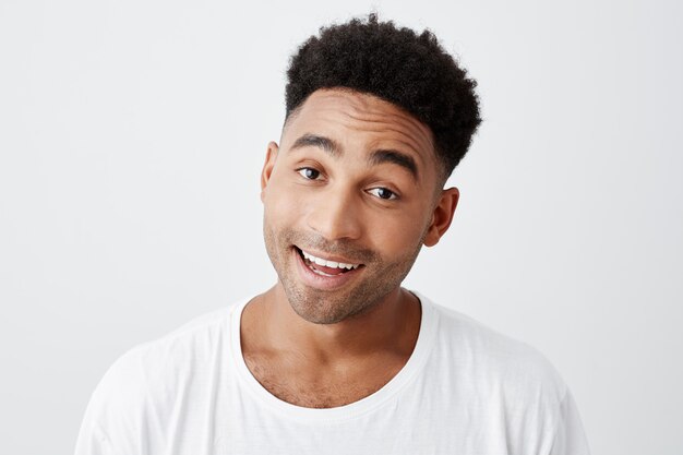 Close up isolated portrait of young beautiful dark-skinned cheerful male student with afro hairstyle in casual white t-shirt smiling with teeth, looking in camera with happy and excited expression.