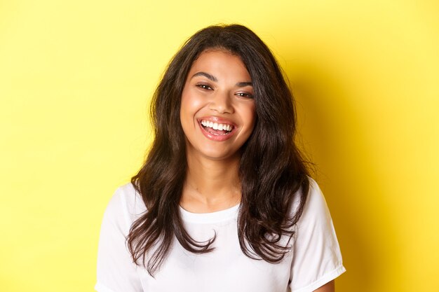 Close-up of joyful and beautiful african-american woman, laughing and looking happy, standing over yellow background.