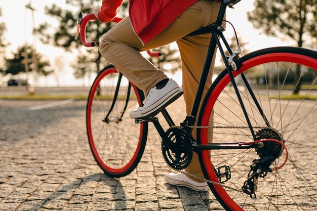 Close up legs in sneakers and hands on steering wheel of hipster style bearded man in red hoodie and beige trousers riding alone with backpack on bicycle healthy active lifestyle traveler backpacker