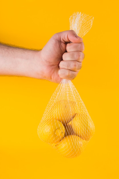 Close-up of a man's hand holding the ripe lemons in the net against yellow background