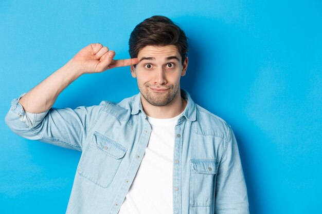 Close-up of man scolding for acting stupid or crazy, rolling finger on head and looking at camera, standing over blue background