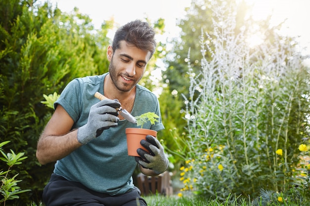 Close up of mature bearded caucasian man in blue t-shirt planting flowers in pot with garden tools,spending peaceful morning in garden hear house.