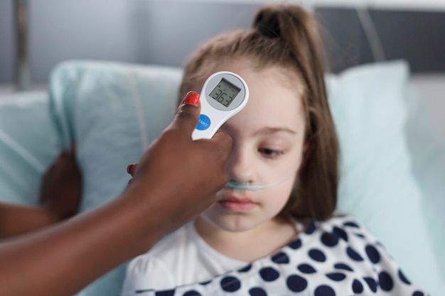 Close up of medical nurse measuring ill little girl body temperature using modern thermometer. Nurse measuring body temperature of sick child while in pediatric clinic recovery ward room.