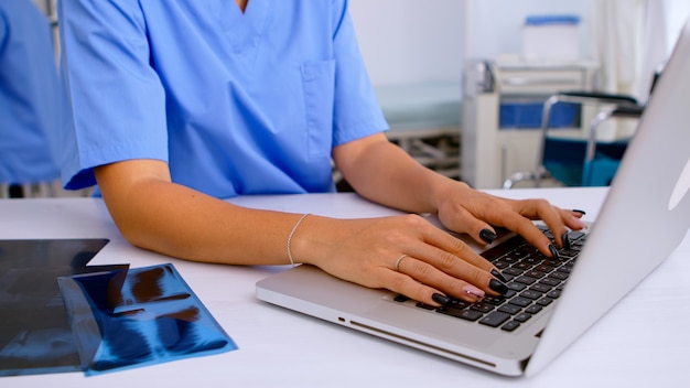 Close up of medical woman nurse writing results of patient radiography, typing on laptop sitting in hospital. Radiologist in medical uniform looking at x-ray, examination, bone, analyze, diagnosis.