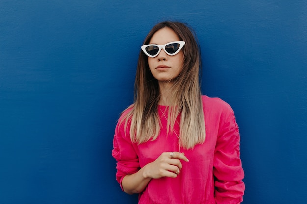 Free photo close up outdoor portrait of charming young woman wearing pink shirt and white sunglasses posing over isolated blue wall