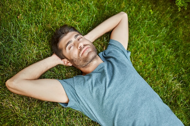 Free photo close up outdoors portrait of young attractive mature bearded hispanic man in blue t shirt looking in camera, lying on ground with relaxed face expression.