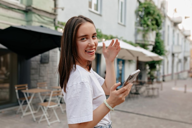 Close up outside photo of charming cute woman with wonderful smile scrolling smartphone and touching her hair on city street