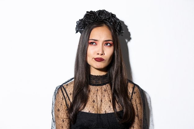 Free photo close-up of perplexed asian woman in wicked witch costume, looking at upper left corner doubtful or indecisive, standing over white background.