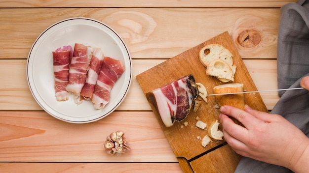 Free Photo close-up of a person cutting the bread slice with rolled up bacon on white dish over the wooden table