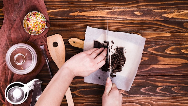 Free photo close-up of a person cutting the chocolate bar with knife on paper over the wooden table