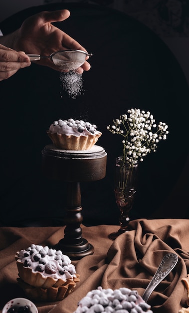 Close-up of a person dusting sugar powder on fruit tart with baby's-breath flowers vase