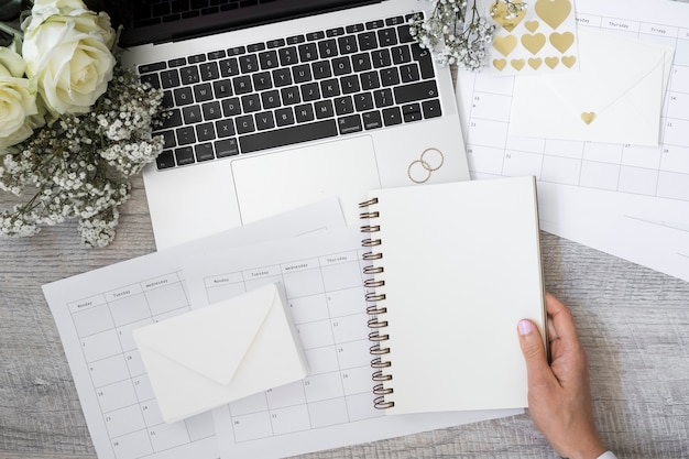 Free photo close-up of a person holding blank spiral notebook with laptop; wedding rings; flower; envelope and calendars on wooden desk