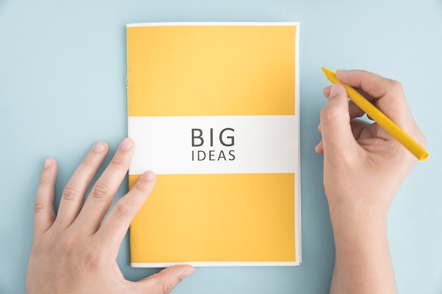 Free Photo close-up of a person holding yellow crayon with big idea book on blue background