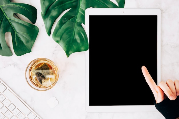 Free photo close-up of a person pointing finger over the digital tablet with tea glass on marble textured backdrop