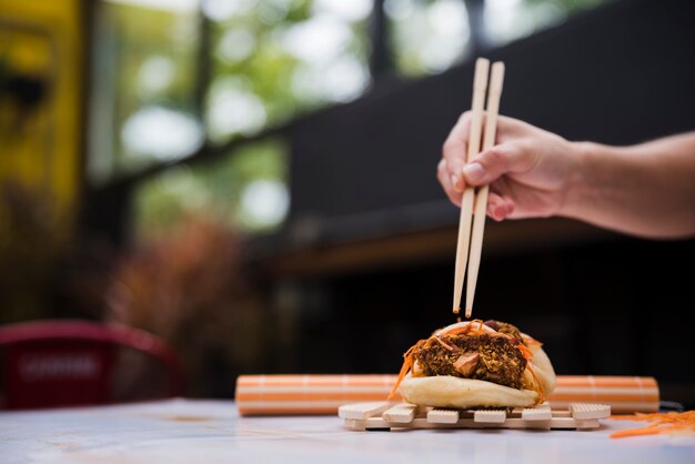 Close-up of a person's hand eating gua bao with chopsticks on wooden tray