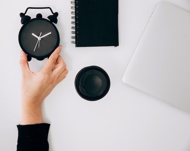 Free Photo close-up of a person's hand holding black alarm clock; spiral notepad; laptop and takeaway coffee cup on white desk